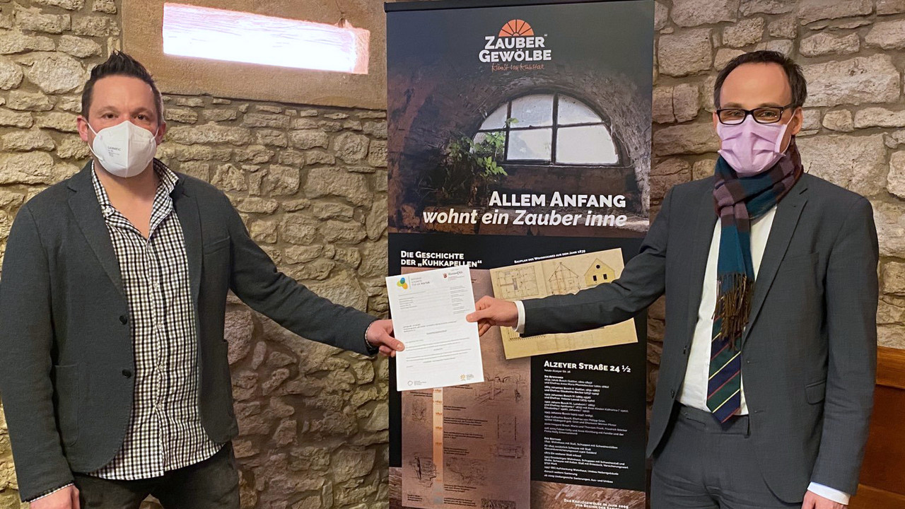 Zwei Männer stehen vor einer Wand und einem Plakat. Der rechte Mann überreicht dem Linken einen Förderbescheid. 