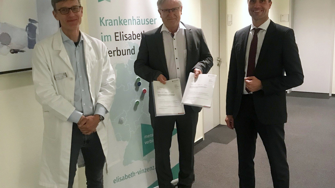 Drei Männer stehen im Gang. Der Mann in der Mitte hält zwei Papiere in der Hand. 