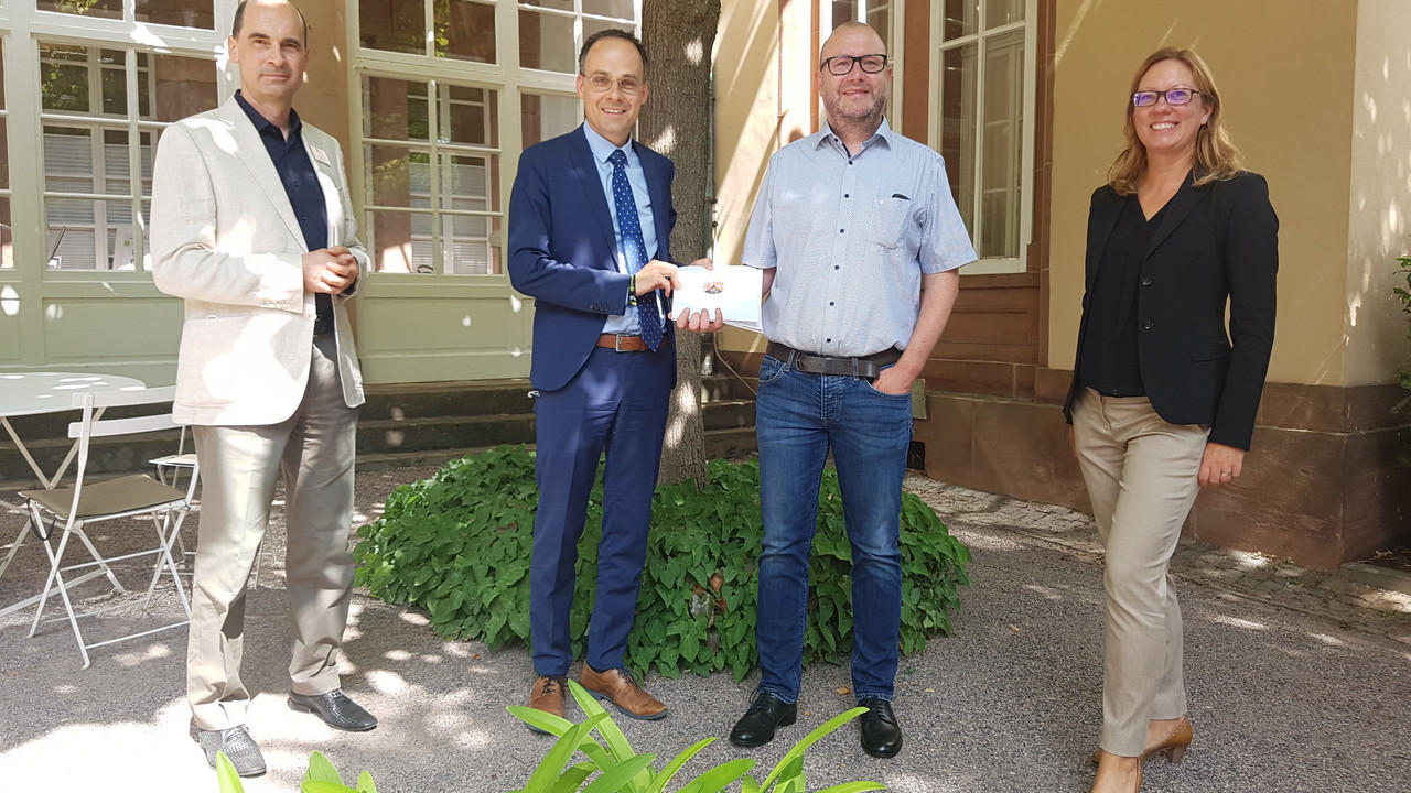 Dr. Denis Alt, Staatssekretär im Ministerium für Wissenschaft und Gesundheit, übergibt Peter Kuhn, Verwaltungsleiter und Leiter der Abteilung EDV, einen Bescheid. Links neben Alt und Kuhn steht Dr. Stefan Wack, Facharzt für Neurologie, Psychiatrie und Psychotherapie. Rechts neben Alt und Kuhn steht Carola Hollnack, koordinierende Gesundheitsreferentin im Ministerium. Das Bild wurde im Außenbereich der Klinik vor einem Baum aufgenommen.