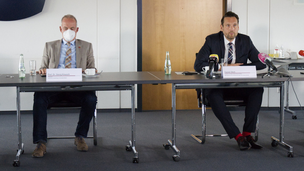 Zwei Männer sitzen am Tisch. Einer trägt eine Maske. Beide sind der Kamer zugewandt. 