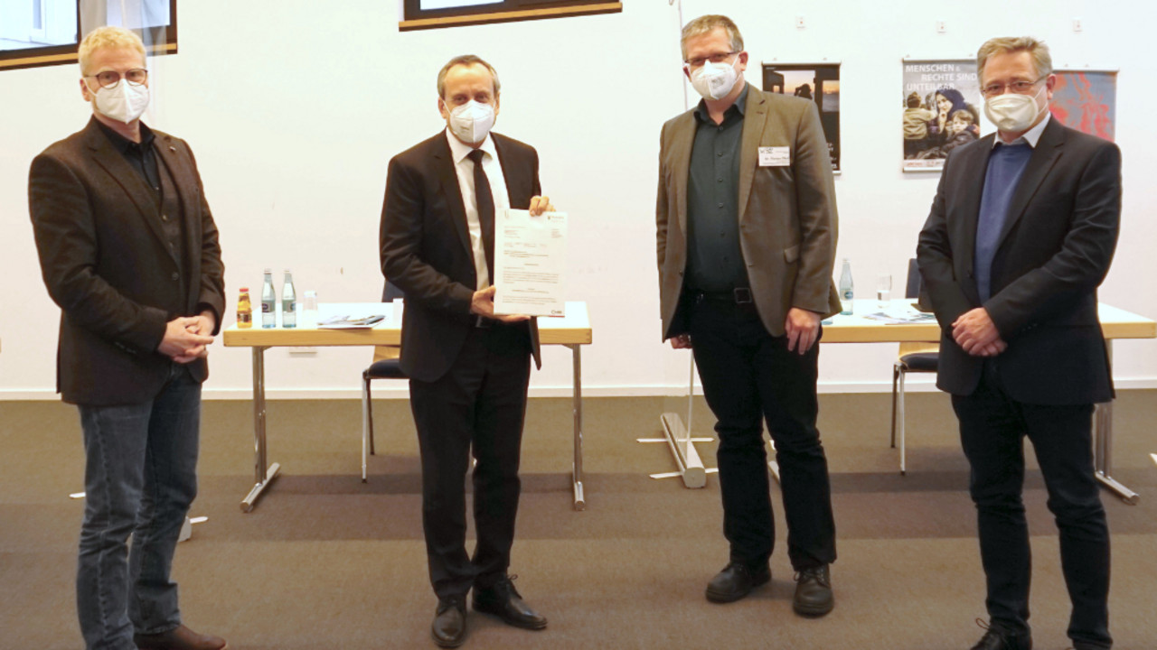 Vier Männer stehen nebeneinander in einem Raum. Der zweite von links hält einen Förderbescheid in der Hand. Alle tragen eine Maske. 